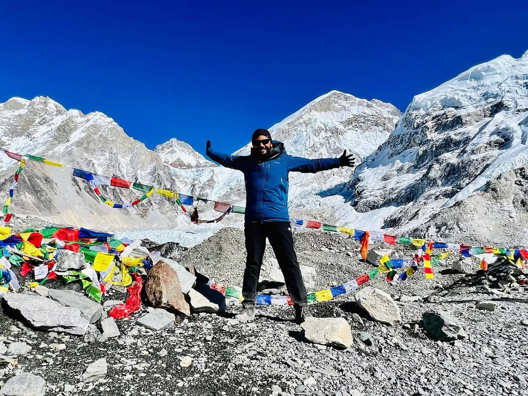 Charith De Silva with arms outstretched at a Himalayan base camp, prayer flags and snow peaks behind him